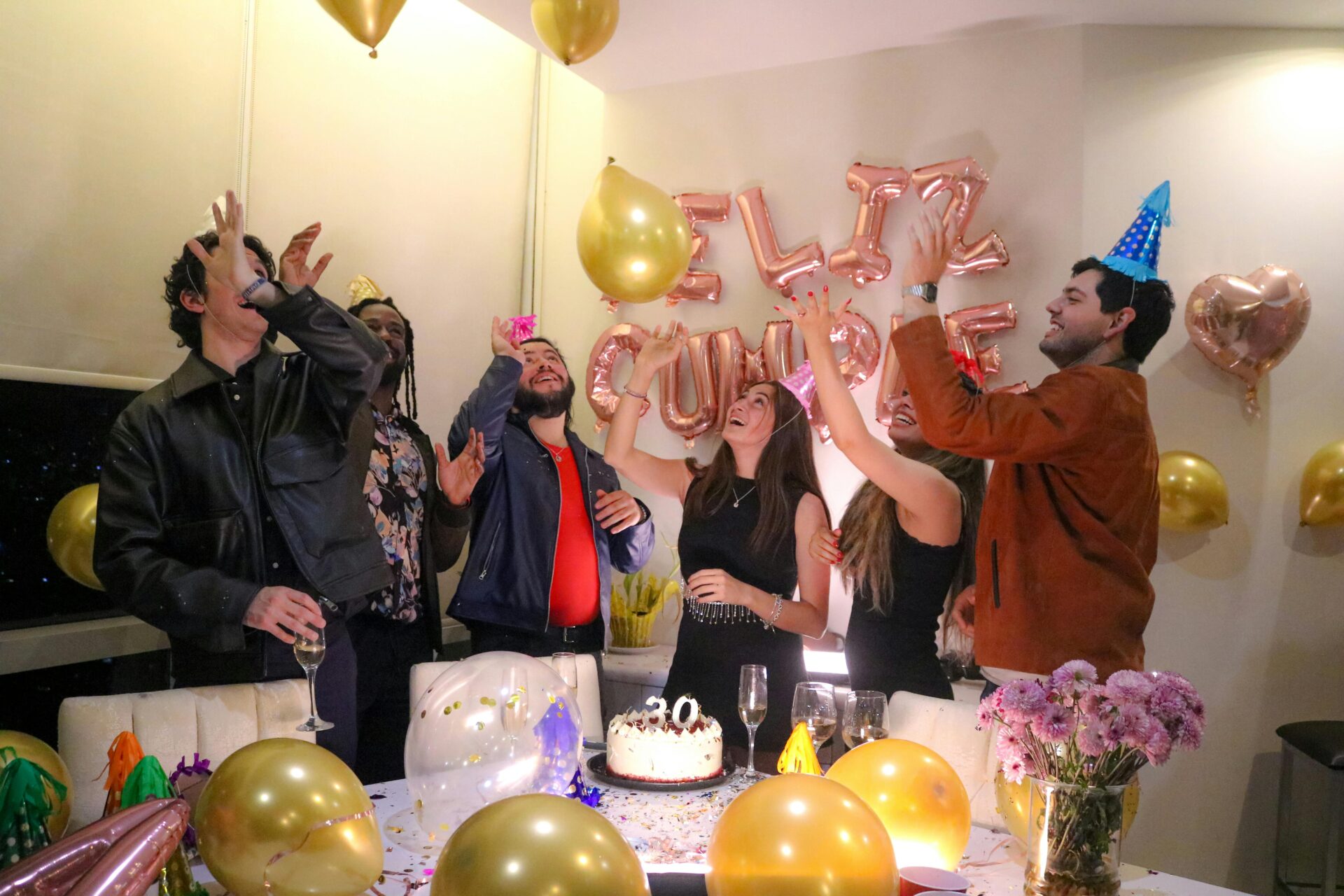 Eine Gruppe von Freunden feiert drinnen einen Geburtstag, umgeben von Luftballons und Kuchen.