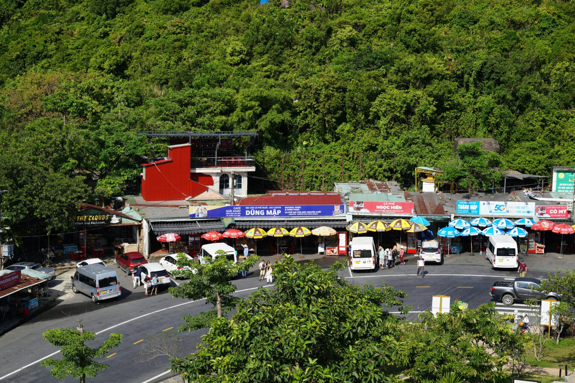 Geschäftiger Straßenmarkt mit bunten Sonnenschirmen und Geschäften, umgeben von üppigem Grün.