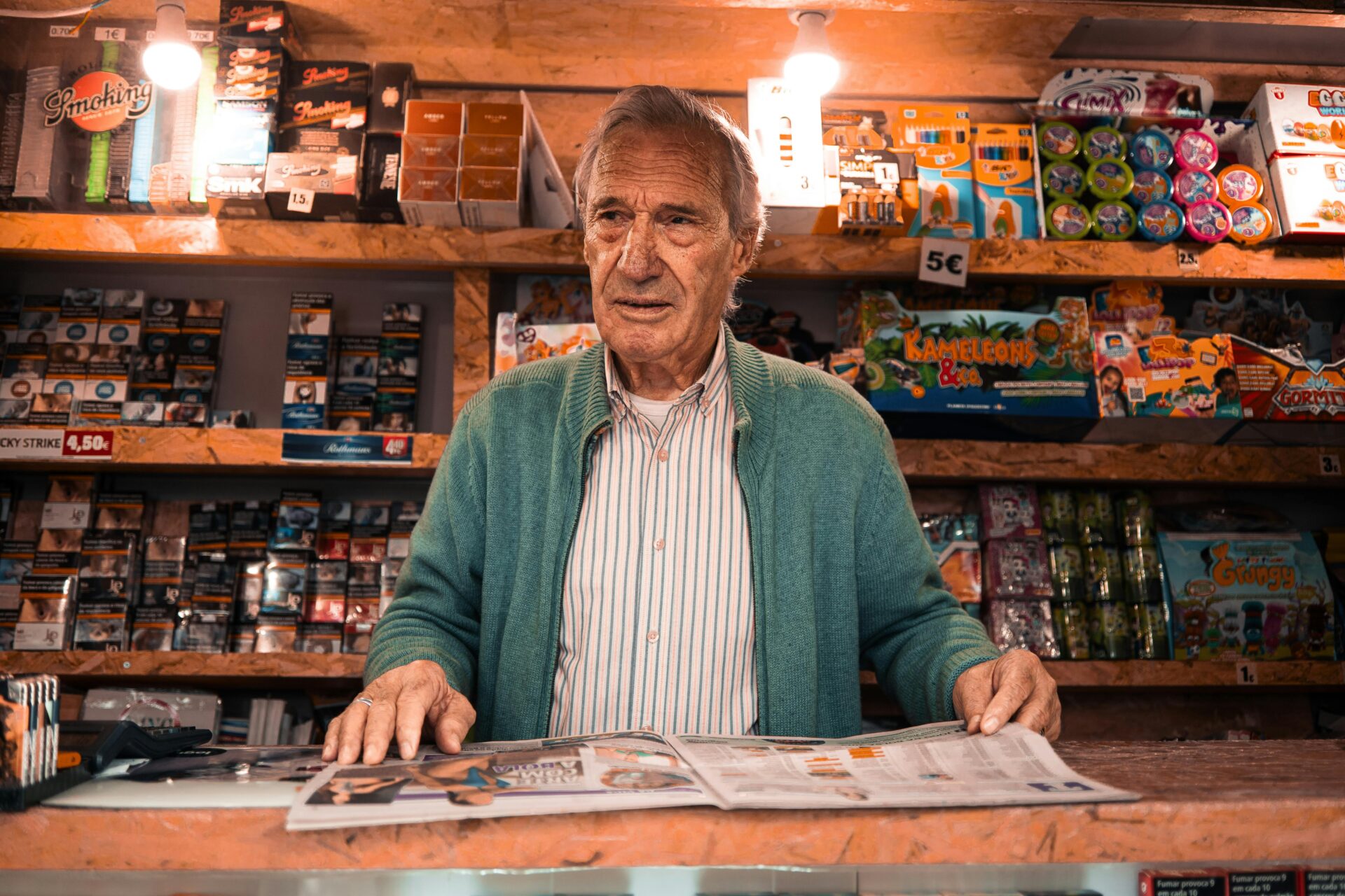 Ein &Atilde;&curren;lterer Ladenbesitzer steht hinter der Theke eines Lissabonner Zeitungskiosks, umgeben von bunten Produkten.