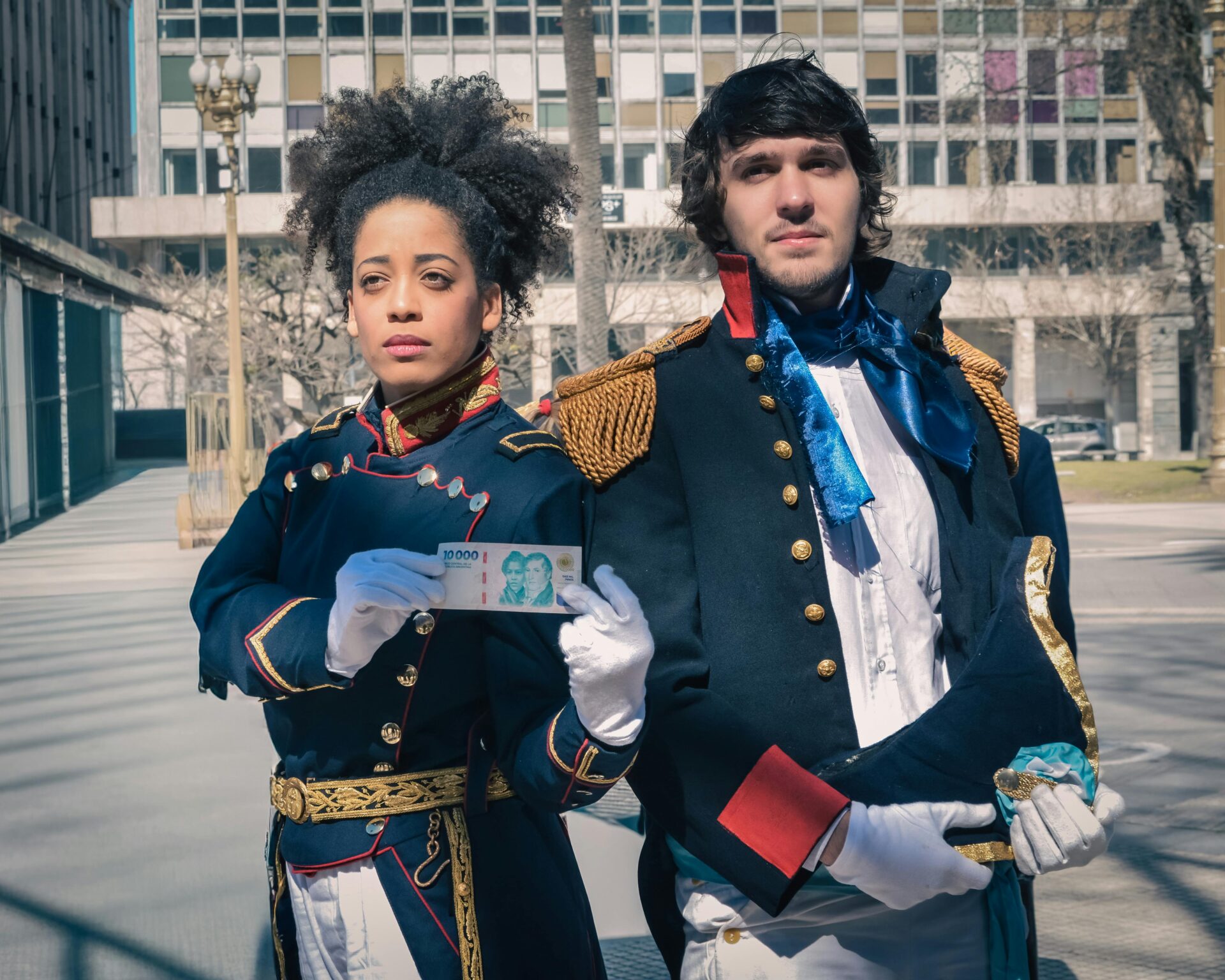 Zwei Erwachsene in historischen Kostümen halten Geld in der Hand, außerhalb einer städtischen Umgebung.